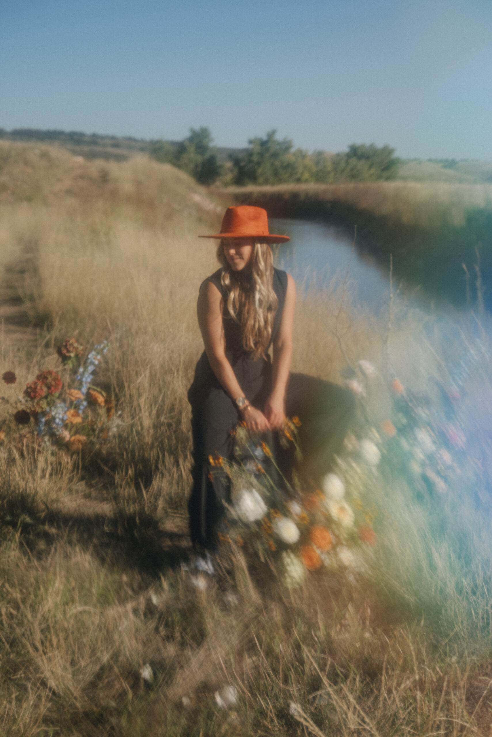 hazy photo of a girl with a hat on sitting on a stool surrounded by floral installments in the middle of a trail near a river with rainbow sun flare in bottom right corner