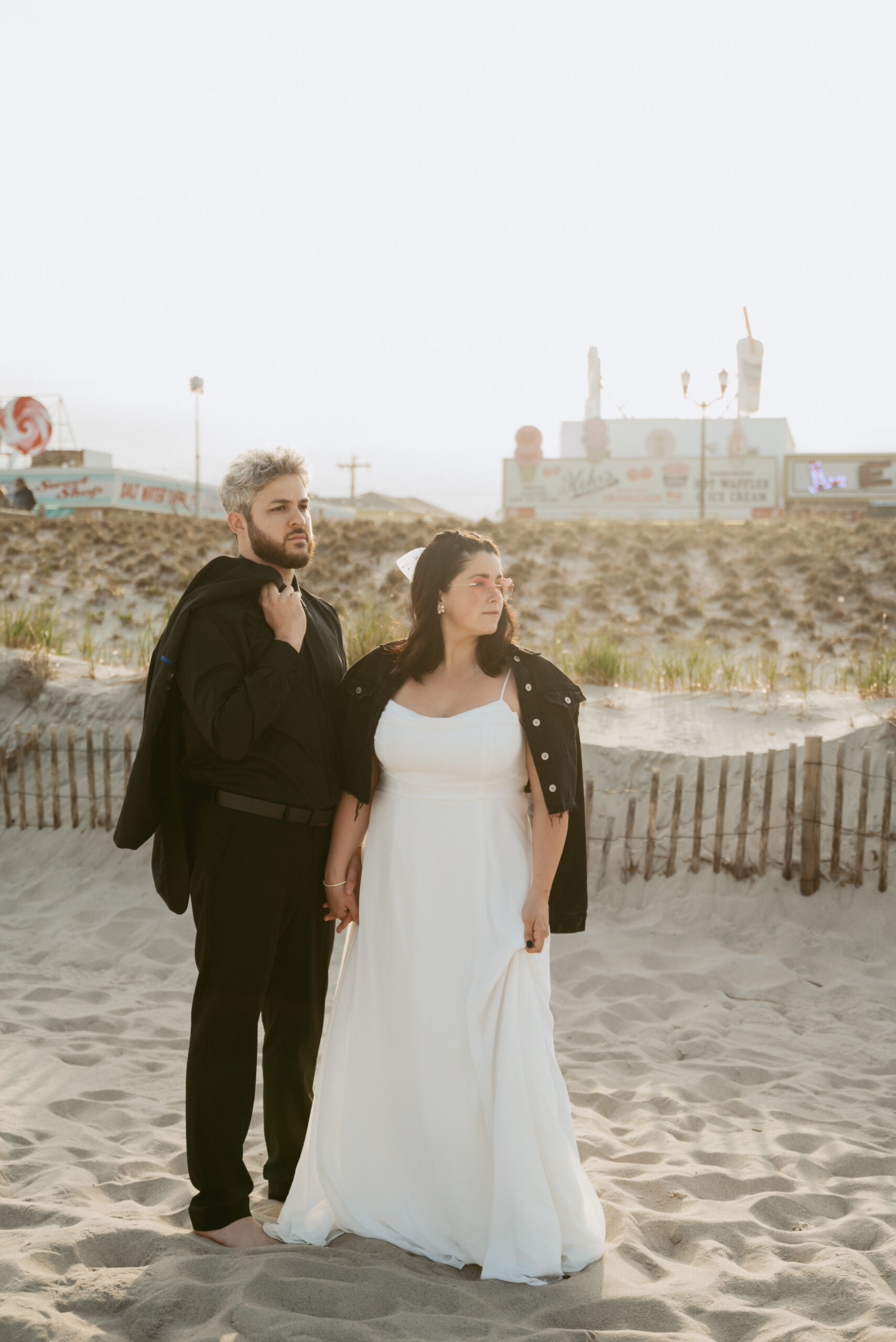 Bride and Groom posing on the beach for wedding photos with arcade and boardwalk behind them. Bride is wearing white dress, heart sunglasses and black jacket on her shoulders and groom is wearing all black with jacket over the shoulder.